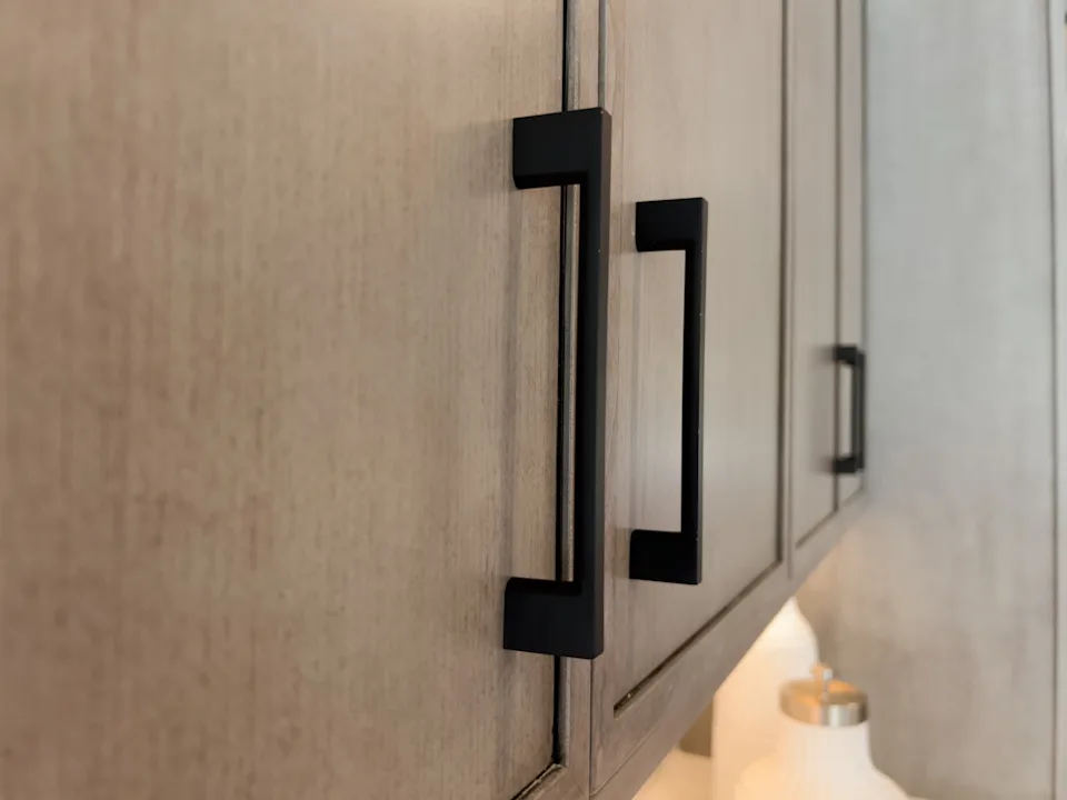 Close-up of a wooden cabinet with sleek, modern black handles. A decorative bottle is partially visible on the countertop below