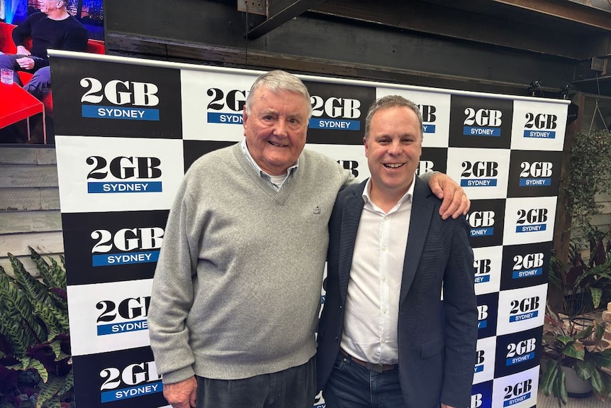 An old man and a younger man stand together in front of 2GB signage in an office