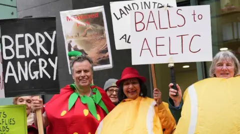 PA Man wearing strawberry costume holding a sign which reads "Berry Angry" and the two women dressed as yellow tennis balls. 