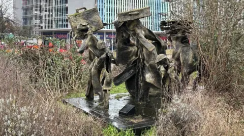 Martin Heath/BBC Close-up of brown and green-coloured sculpture which features a group of people. The two adults at the front are carrying suitcases on a shoulder and on the head. The sculpture is surrounded by grass and other flora.
