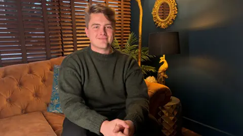 Elliott Cockett pictured with short blonde hair smiling at the camera. He is wearing a dark green sweater.