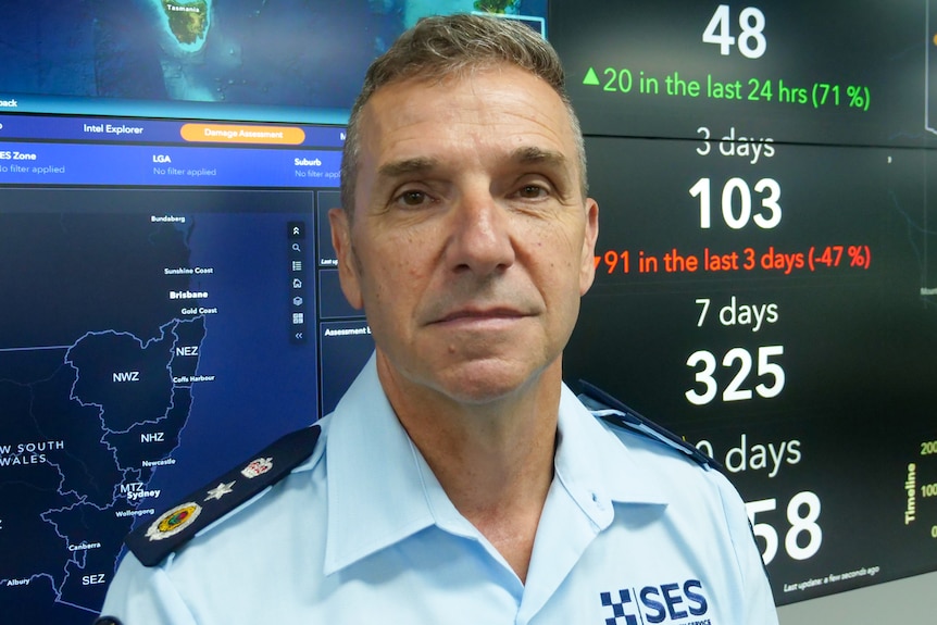 A middle-aged man in an SES uniform stands in front of a bank of screens displaying maps and data.