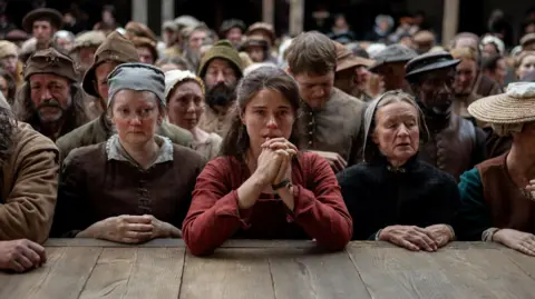 Universal Pictures/Focus Features Jessie Buckley hair long brown hair draped behind her hair. She has a said facial expression with her hands clasped together in front of her, with her elbows resting on a wooden stage. She is wearing a red long sleeved dress. She is in the middle of a large crowd of men and women in period clothing. 
