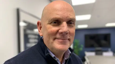 Jim is smiling at the camera. He is standing in a room with navy walls and there is a large meeting table and chairs in the the centre. A flat screen TV is hanging behind him. He is wearing a purple shirt and navy quarter zip jumper.