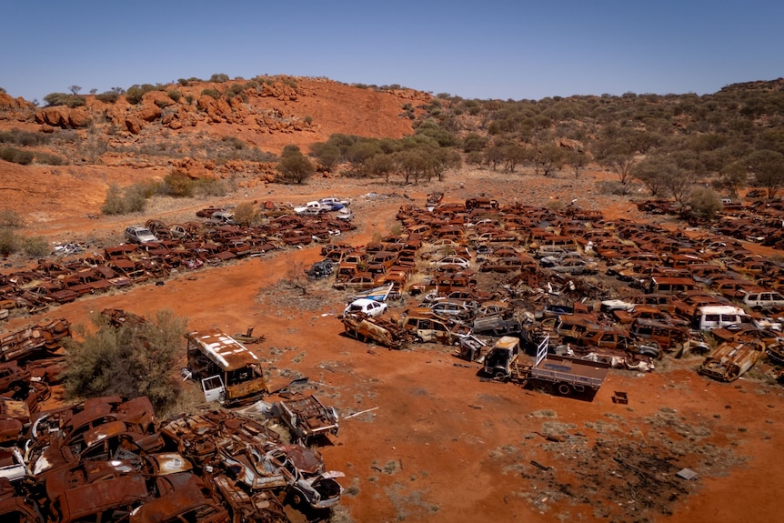 Piles of rusted cars on similar colour rusty sand and ochre hills behind it. 