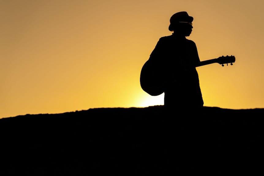 A sillouhette of Desmond wearing a hat. The yellow sun behind him. He has a guitar in his hand. 