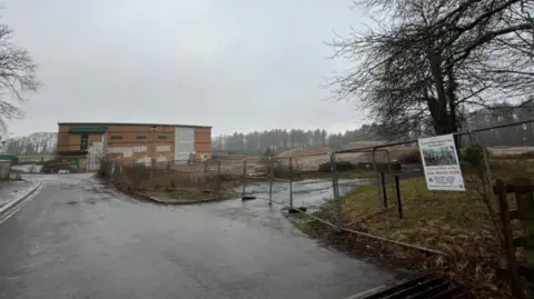 A derelict school in the Forest of Dean, surrounded by woodland on a misty winter's day, and metal construction fencing, with signs on it. There is a road leading up to the school, a light brick building with green and white windows and cladding.