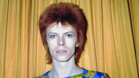 Getty Images An image of David Bowie from the 1970s, he is standing against a yellow curtain and has dark red hair. 