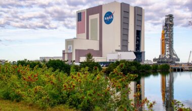 An orange rocket sits on a mobile scaffold as it rolls out of a large white building with the American flag and NASA meatball logo on the side.