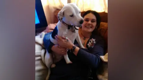 Sam Cook Paula Parretti sits on a sofa with a white dog on her lap. She is holding the dog around its chest and she is smiling at the camera. She is wearing a dark coloured long-sleeved top with a floral pattern on her left chest, and is also wearing a black necklace.