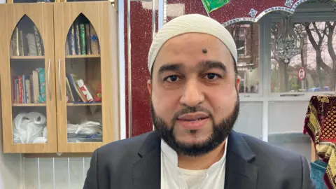 Cathy Minton/BBC Mohammed Ilyas is pictured in the mosque. He has dark hair and a short beard and is wearing a dark suit jacket over a white shirt and white T-shirt.