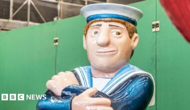 A sailor sculpture in the style of Beryl Cook's paintings sits with his arms crossed. He's wearing a white hat and is smiling.