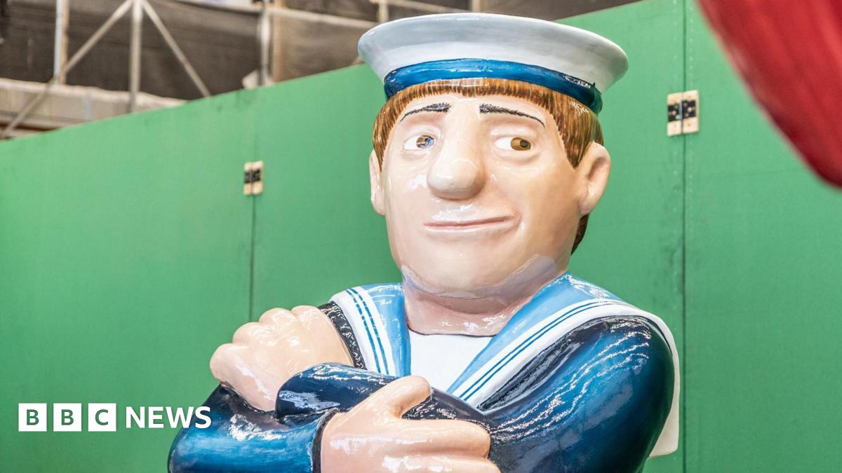 A sailor sculpture in the style of Beryl Cook's paintings sits with his arms crossed. He's wearing a white hat and is smiling.
