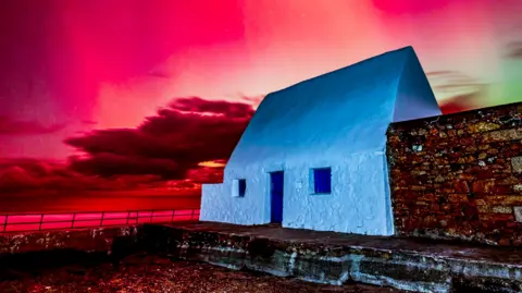 MONSTERCURRYMAN/BBC WEATHER WATCHERS white cottage with a blue door. the sky is red and green due to the Northern Lights 