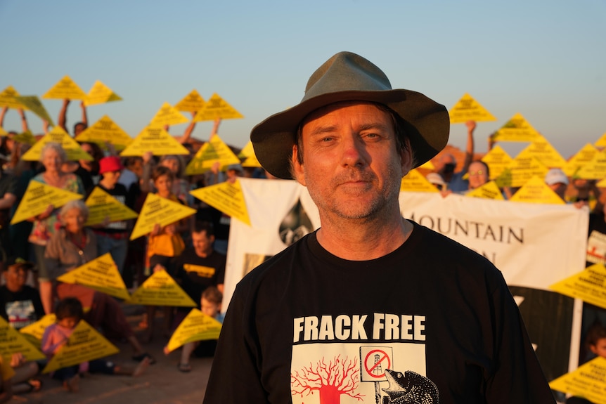 a man in a hat smiling at the camera, with people holding placards behind him 
