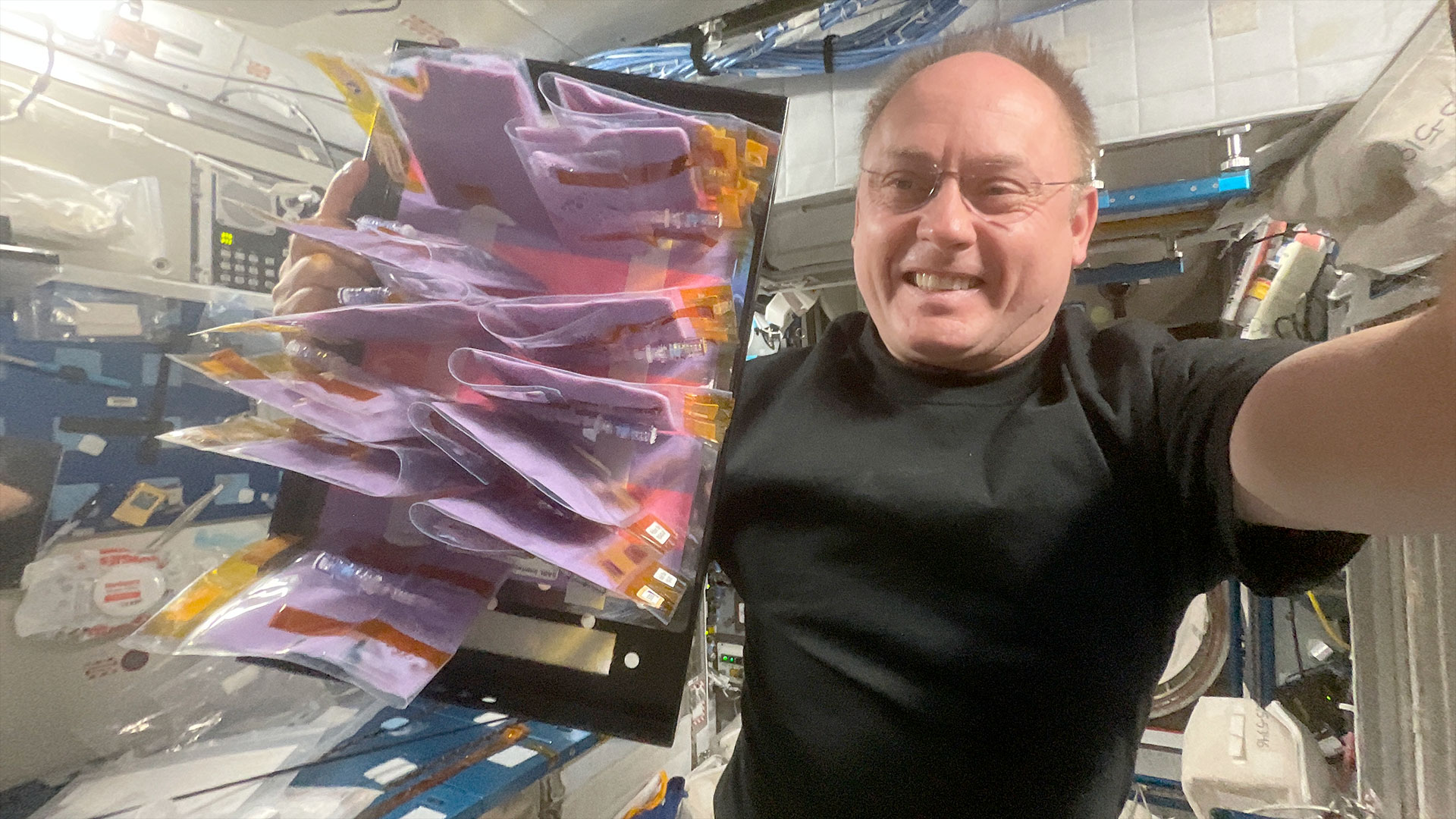 a man in a black t-shirt holds up a tray of plastic pouches filled with purple fermented milk aboard a space station.