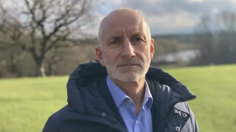 Alistair Smith has short grey hair and stubble. He is wearing a warm padded coat and a light blue shirt underneath. He is stood in a field with trees in the background.