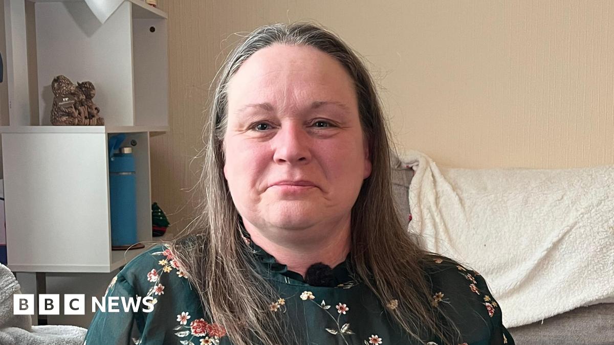 Elaine is looking at the camera. She has long brown/grey hair and is wearing a green floral top with a high neckline.