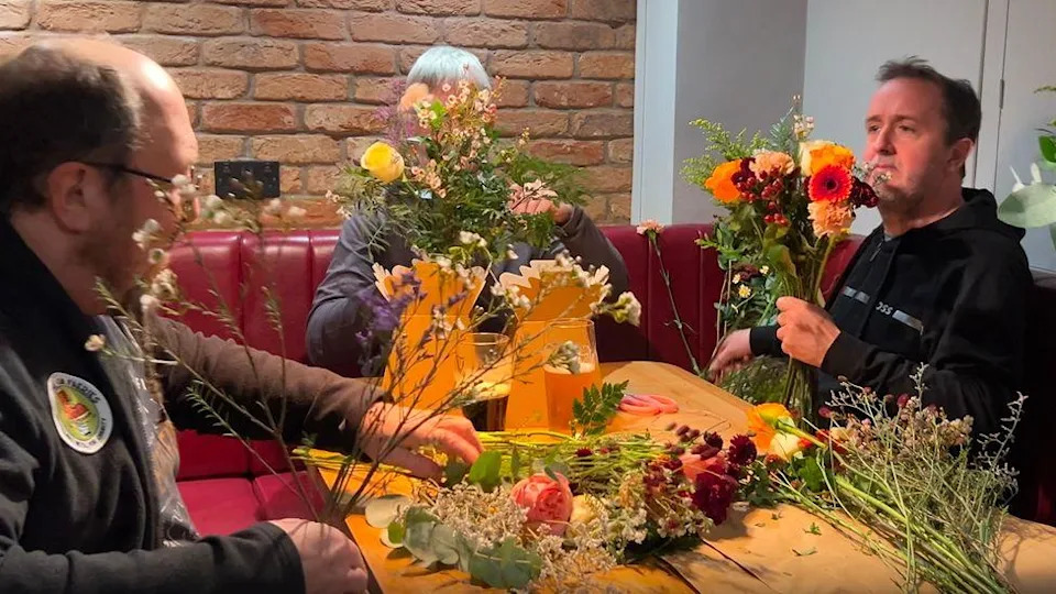 Three men sit round a pub table holding flowers that they are arranging in their hands. On the table are more flowers and pints of beer.