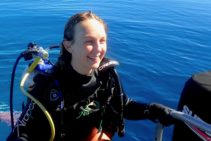 A young woman in scuba gear climbing onto a boat from its back ladder out of the blue ocean.