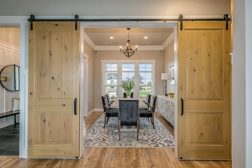 Open sliding barn doors reveal a dining room with a table, chairs, chandelier, and a large window overlooking the outside