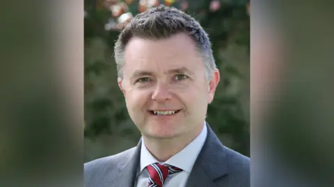 Royal School Armagh Graham Montgomery smiling at the camera. He is a man with short greying hair.  He is wearing a grey blazer, a pale shirt, and red tie with white and navy stripes.