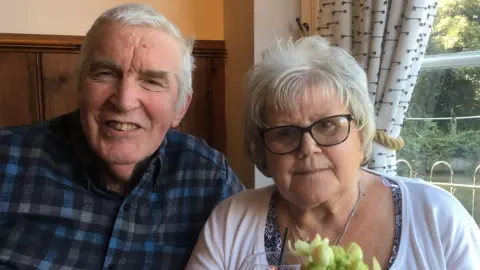 Ralph Taylor Ralph and Carol Taylor are sat down at a table and looking at the camera. Ralph is on the left of the image. He is wearing a dark chequered open-neck shirt and is sat in front of wood panelling. Carol is wearing a white cardigan over a floral-patterned top and sits in front of a pulled-back white curtain. They both have grey hair, and Carol wears black-rimmed spectacles.