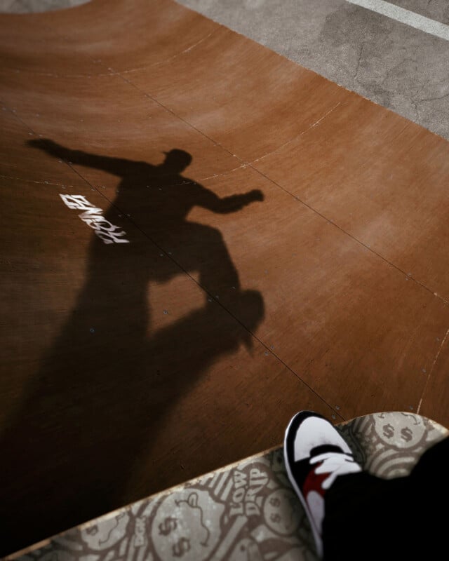 A shadow of a skateboarder performing a trick on a brown ramp, with part of a person’s shoe and patterned seat visible in the lower right corner.
