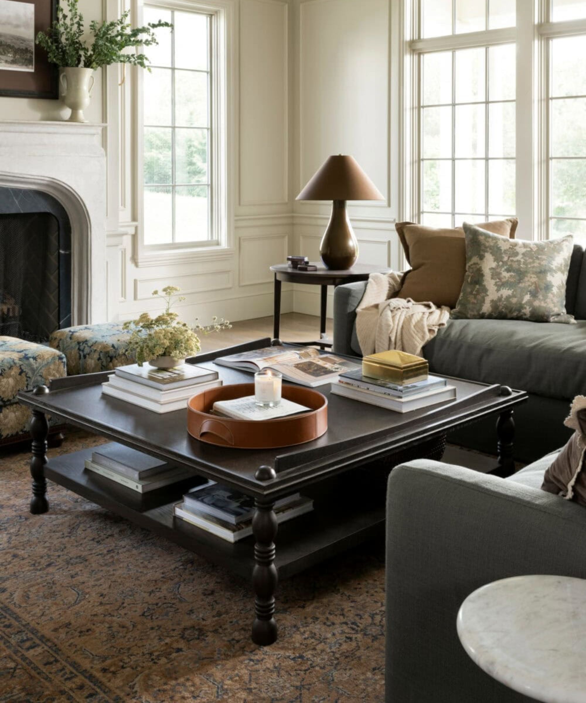 neutral living room with paneled walls, large windows and doors, a large corner sofa in dark grey with a black coffee table styled with books, foliage in a vase, a tray with a candle