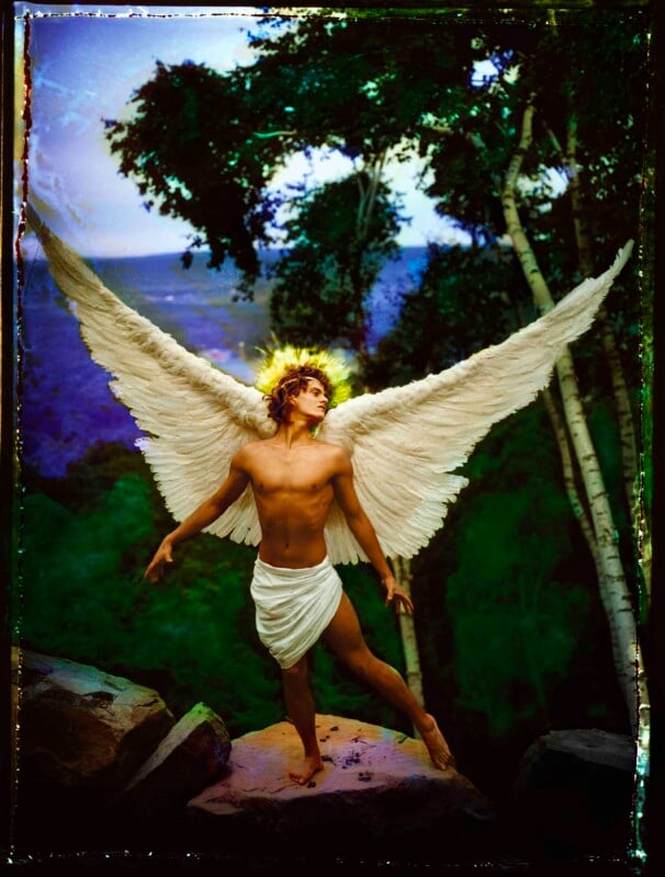 A shirtless figure with large white wings and a glowing halo stands on rocks in a forest, wearing a white cloth around the waist. Tall trees and a mountain are visible in the background.