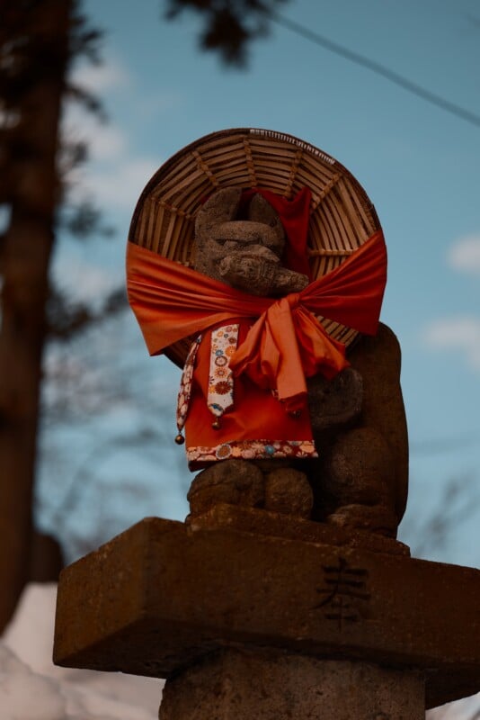 Stone statue of a fox wearing a red cloth and a woven hat, standing on a pedestal with Japanese characters. The sky is blue with scattered clouds, and there are blurred trees in the background.