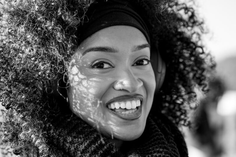 Smiling person with curly hair, wearing a headband and scarf, listens to headphones. The sun casts dappled light across their face. Black and white image.