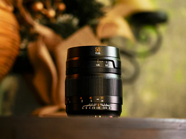A black 50mm camera lens sits upright on a dark surface, with a softly blurred background featuring warm tones and hints of green and brown decor.