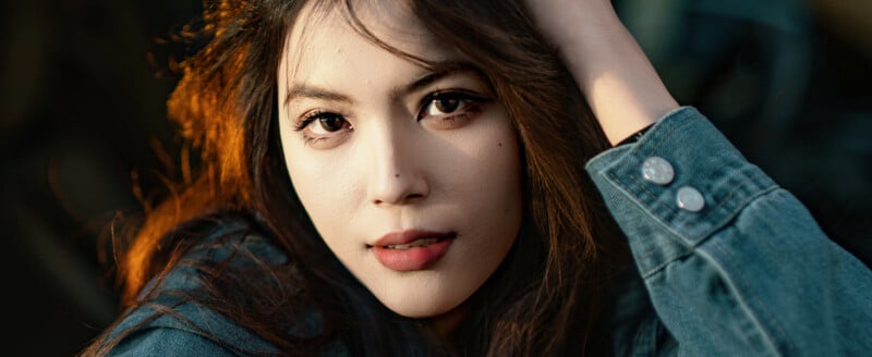 Close-up of a young woman with long brown hair wearing a denim jacket, gazing directly at the camera with a relaxed expression and one hand touching her head, in warm natural light.