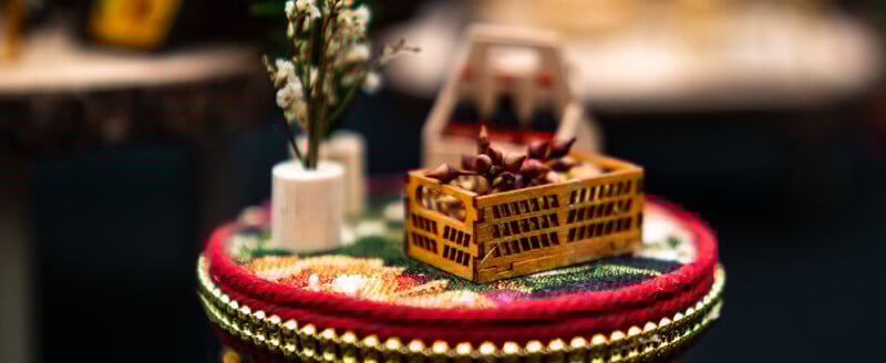 A miniature wooden crate with small red fruits sits on a colorful, decorative round table alongside a tiny potted white flower and a small bottle carrier in the background.