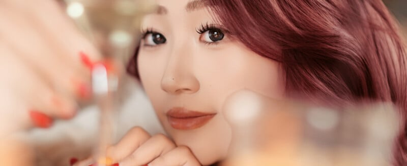 A close-up of a woman with long, wavy, reddish hair and natural makeup, looking at the camera while holding a glass. There are soft, warm lights creating a cozy and festive atmosphere.