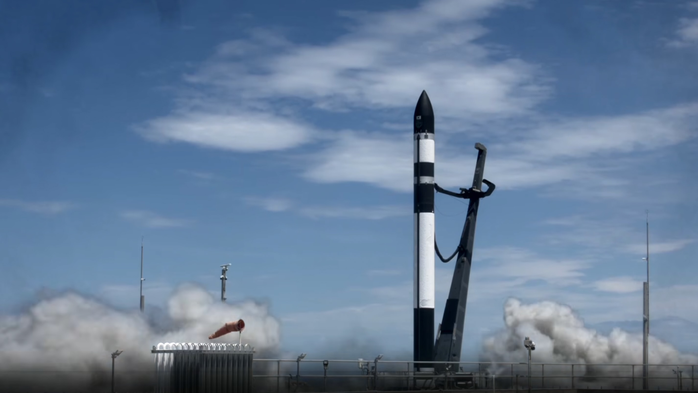 a black and white rocket stands on a launch pad beneath a blue sky, with whitish-gray exhaust billowing from its base