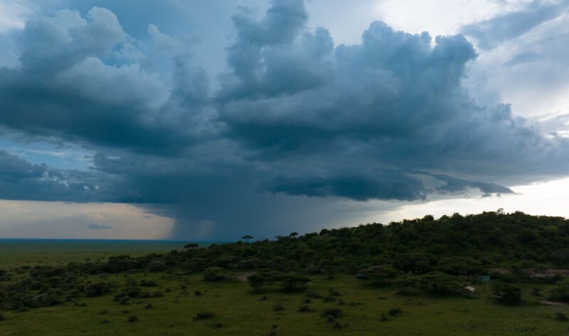Dark storm clouds gather over a green savanna landscape, with rain visibly falling in the distance and a hill covered in trees in the foreground. The scene is dramatic and atmospheric.