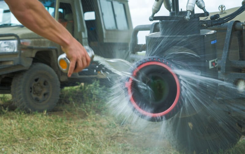A person sprays water onto the lens of a large camera rig outdoors, causing water to splash. A vehicle is parked in the background on grassy terrain.