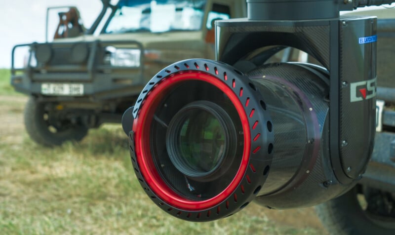 A close-up view of a large camera lens with a red ring, mounted on a stabilizing rig outdoors, with a blurred off-road vehicle in the background on grassy terrain.