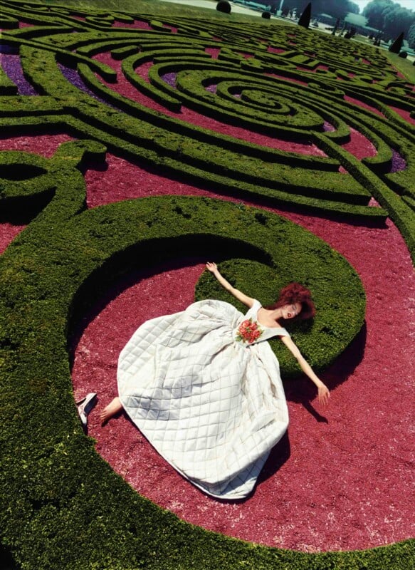A woman in a voluminous white dress lies with arms outstretched on red gravel within a green, circular hedge maze in a formal garden. One shoe has fallen off, and she looks relaxed and dramatic.