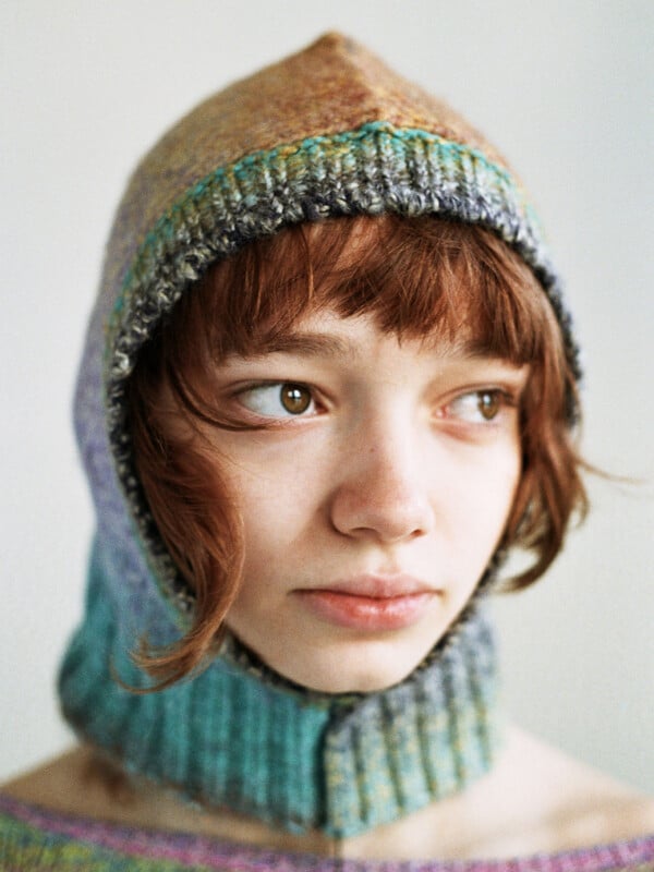 A young person with light skin and short, wavy brown hair looks slightly to the side while wearing a colorful, knitted hood that covers the head and neck, with soft natural lighting in the background.