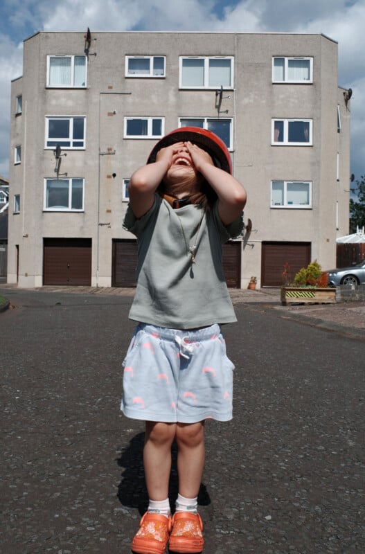 A young child stands on a paved street in front of a grey apartment building, wearing an orange hat, grey shirt, white shorts with pink patterns, and orange shoes, covering their face with their hands.