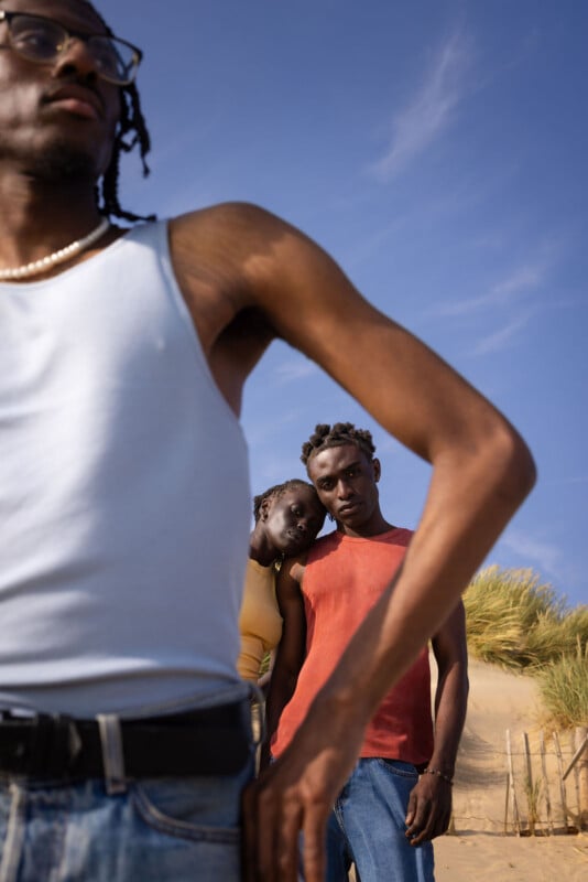 Three people stand outside on a sandy dune under a blue sky. One person in a tank top is in the front, partially out of frame. Two others stand close together behind, with one resting their head on the other's shoulder.