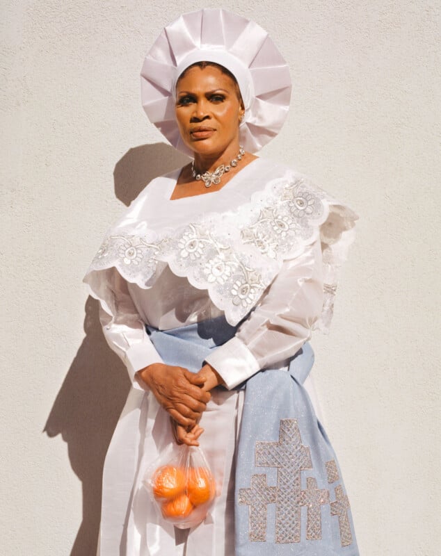 A woman stands against a light wall wearing a white lace dress, a matching headpiece, a blue apron with silver cross designs, and holds a small bag of oranges in one hand.