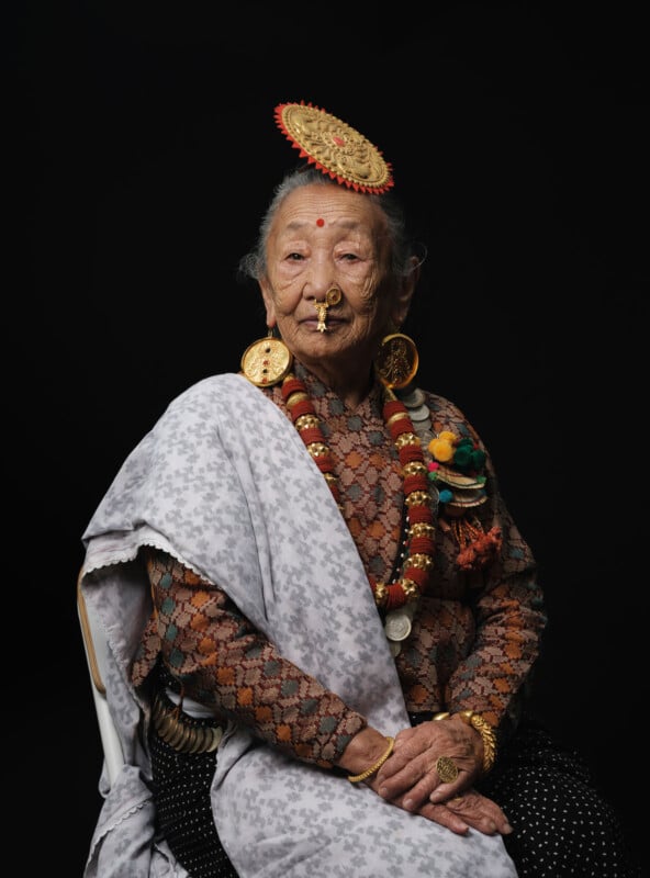 An elderly woman with long gray hair sits against a black background, wearing traditional jewelry, a patterned shawl, and a decorative circular headdress. She looks calmly at the camera.