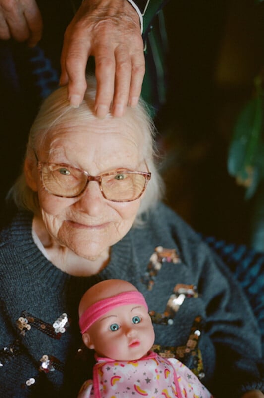 An elderly woman with glasses smiles warmly, wearing a sequined sweater. A hand rests gently on her head, and she holds a baby doll dressed in pink clothing and a headband.