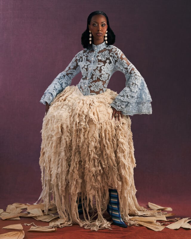 A woman stands confidently against a dark backdrop, wearing a blue lace top with flared sleeves, a voluminous beige textured skirt, striped blue boots, large pearl earrings, and corn husks scattered on the floor.