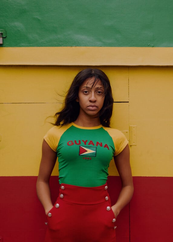 A woman with long dark hair stands against a red, yellow, and green wall. She wears a green shirt that says "GUYANA" with the country's flag and red high-waisted pants. She looks directly at the camera with a neutral expression.