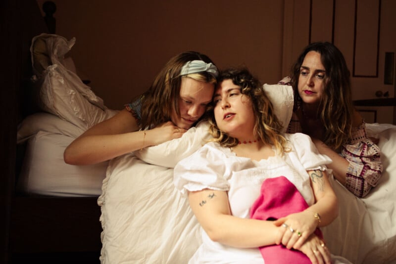 Three women sit closely together on a bed, appearing thoughtful and relaxed. Two lean on pillows while one sits in front, wearing a white dress with a pink section. Warm, soft lighting fills the cozy room.